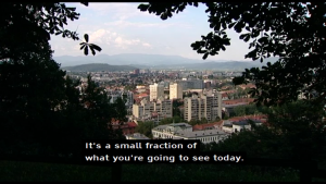 A screen shot from a broadcast, displaying some buildings, and machine-translated subtitles in English, which states "It's a small fraction of what you're going to see today."