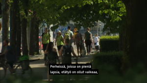 View of a summer day in a park and several people walking in the shade of large trees, with the Finnish machine-translated subtitle "Perheissä, joissa on pieniä lapsia, etätyö on ollut vaikeaa," ("Working from home has been difficult for families with small children,")